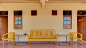A room with a wooden bench, two yellow chairs, and a small table. There are three windows with blinds, and a wooden door on the right. - Hotel O Wisma Subur Syariah (Pekalongan)