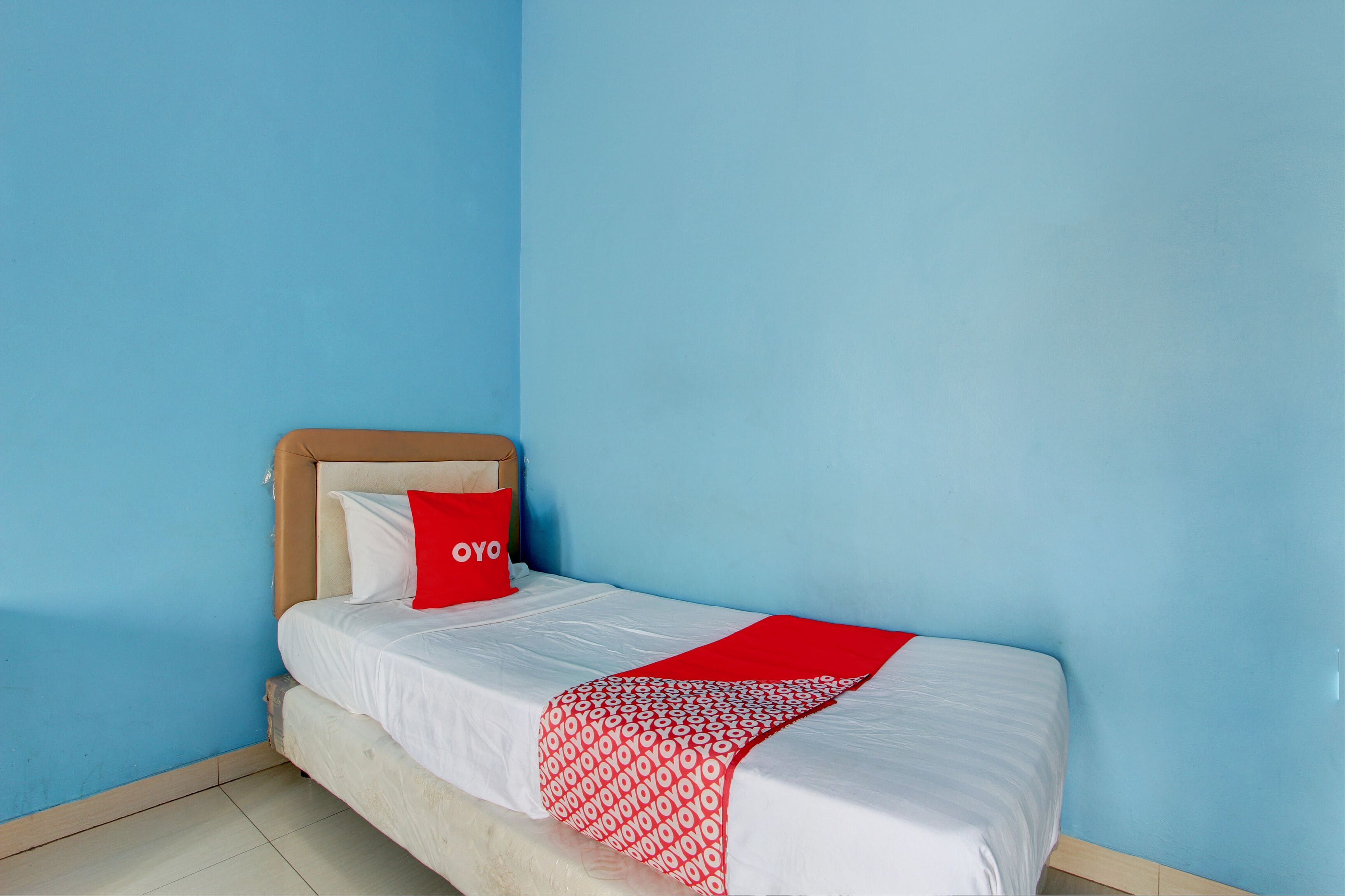Un lit simple avec un oreiller rouge et une couverture à motifs, dans une pièce aux murs bleus et dotée d’une tête de lit en bois.
