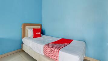 Un lit simple avec un oreiller rouge et une couverture à motifs, dans une pièce aux murs bleus et dotée d’une tête de lit en bois.