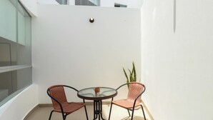 Un petit balcon avec une table ronde en verre et deux chaises en osier, une plante en pot et un mur blanc.