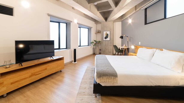 A modern bedroom with a large bed, a wooden TV stand, and a large window providing natural light. - w* | Cozy Loft in Laureles (Medellín)