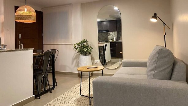 Un salón moderno con un sofá gris, una zona de comedor con una mesita y sillas, una cocina al fondo y un espejo grande en la pared.