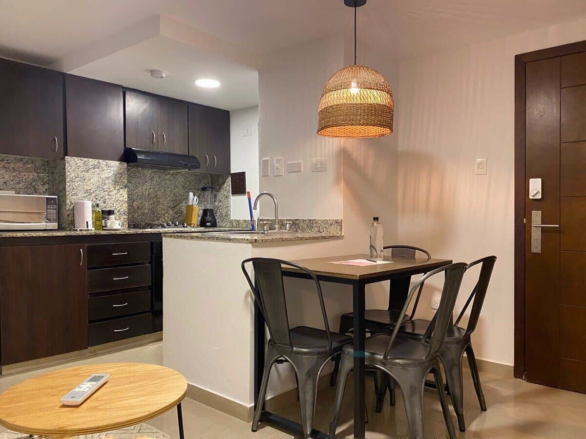 A compact kitchen with dark cabinets, a granite countertop, and a central island with a sink. There is a dining area with a table and chairs, and a hanging pendant light above the island.