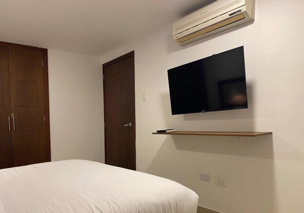 Un dormitorio con una cama, un televisor montado en la pared y un aire acondicionado sobre la cama.