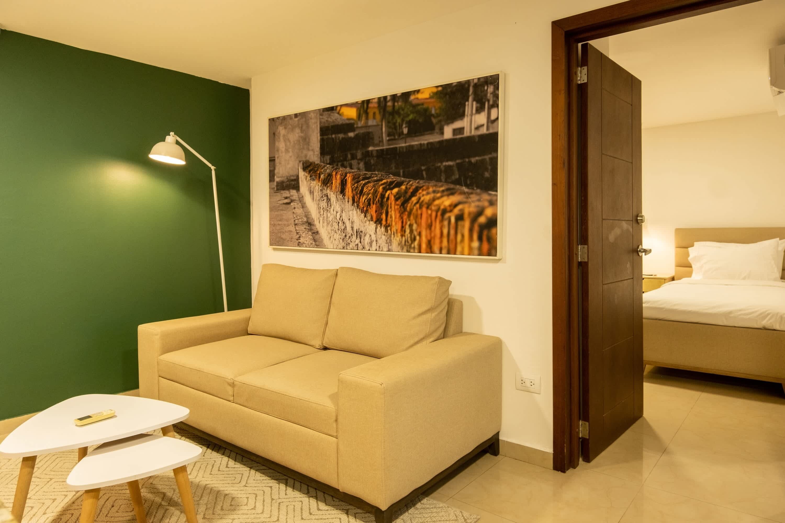Une pièce avec un canapé beige, une petite table blanche, un lampadaire et un lit avec une literie blanche.