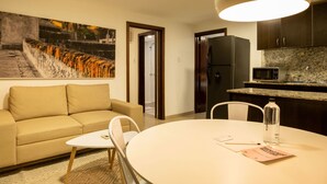 Ein modernes Wohnzimmer mit einem beigefarbenen Sofa, einem runden Esstisch und einer Küchenzeile mit dunklen Schränken und einer Mikrowelle.