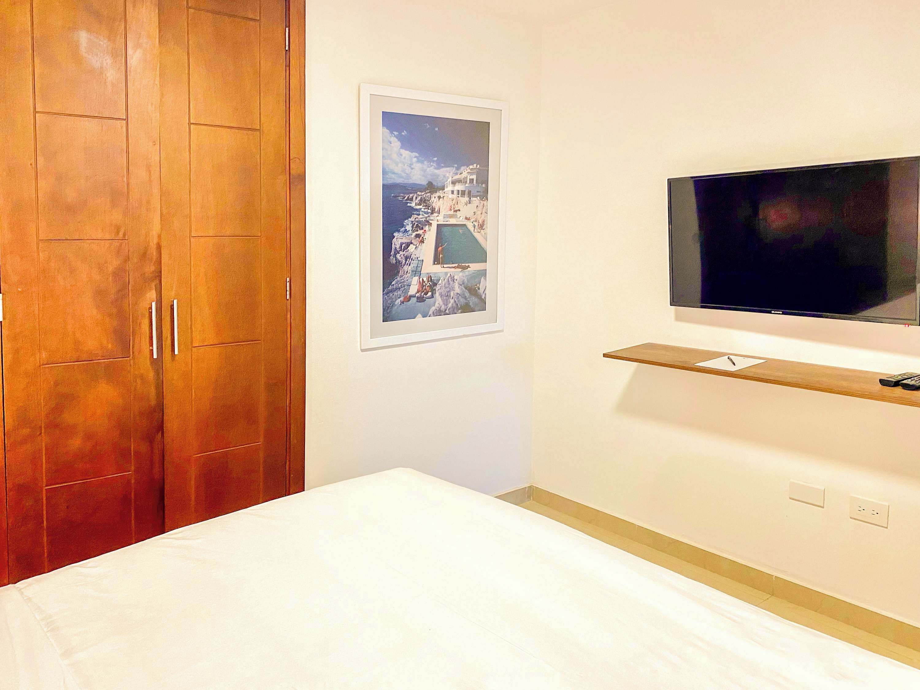 A bedroom with a wooden wardrobe, a framed picture on the wall, a television mounted on the wall, and a bedside table with a remote control.