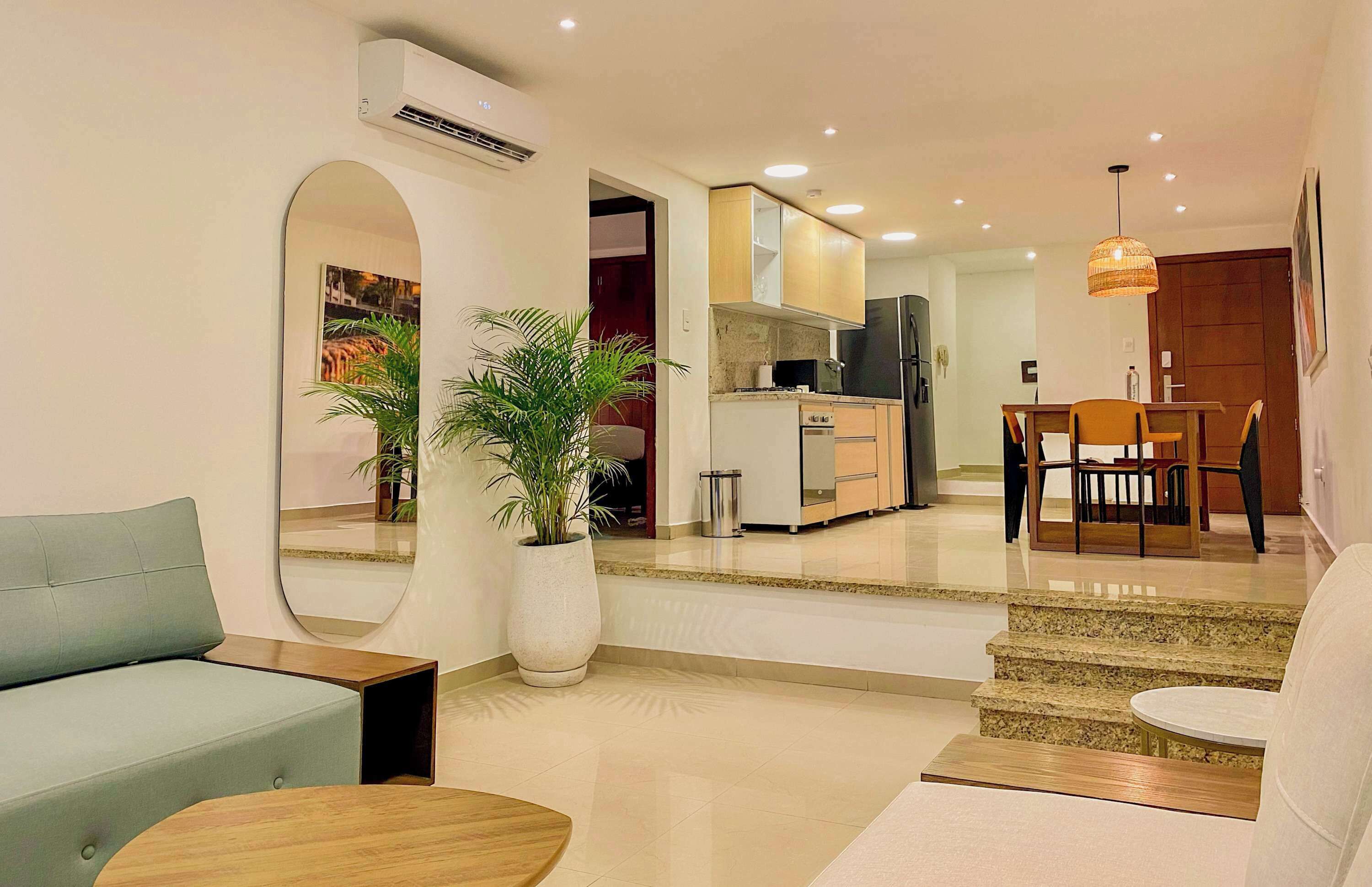 Una sala de estar moderna con un sofá, una mesa de centro y un comedor con mesa y sillas. Se observa una cocina al fondo con un refrigerador y un microondas.