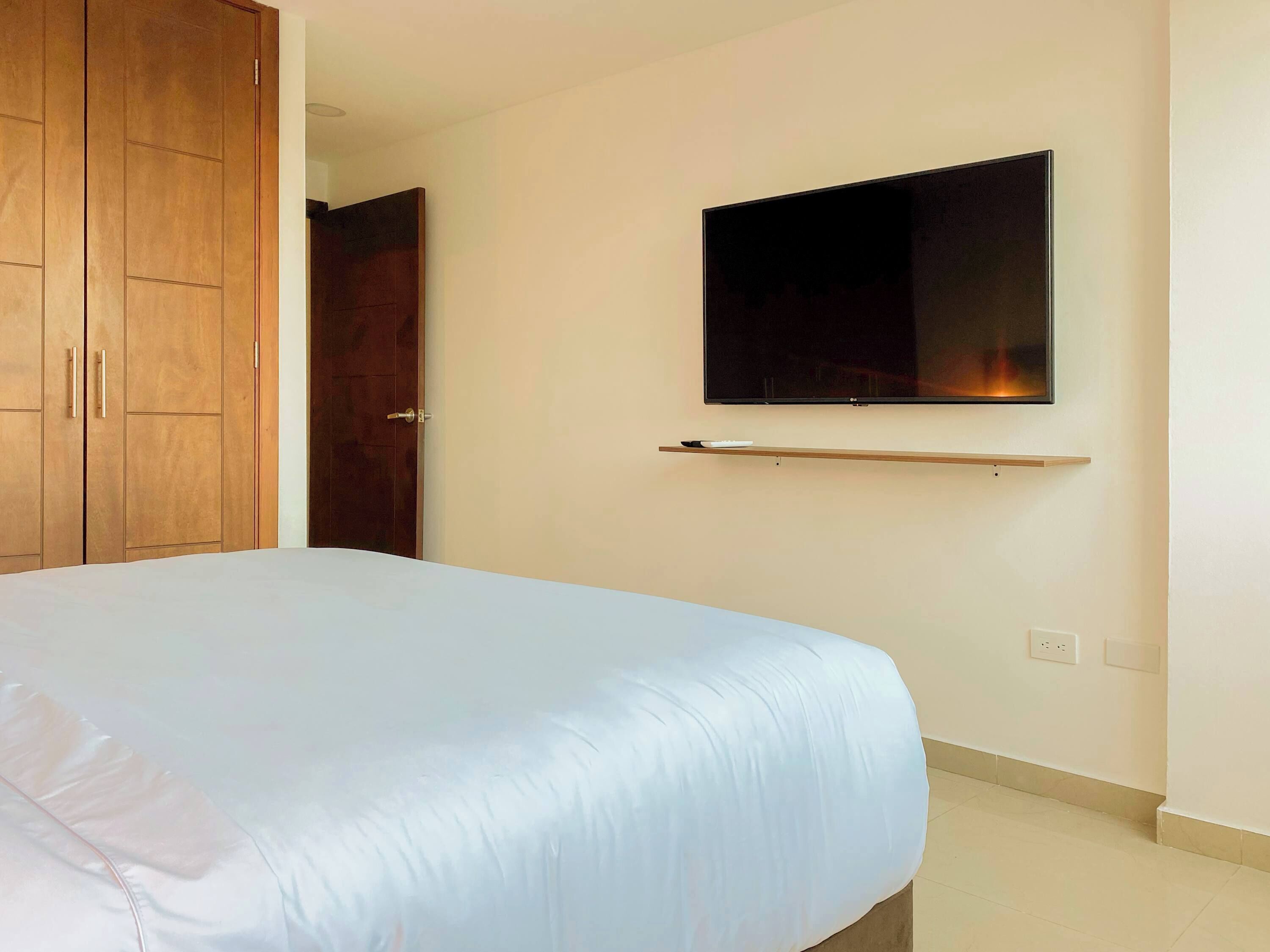 A bedroom with a bed, a wardrobe, a television mounted on the wall, and a small shelf below the TV.
