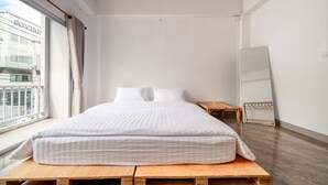 Quarto minimalista com uma cama grande, uma mesinha de cabeceira de madeira, um espelho de corpo inteiro e vista para um edifício.