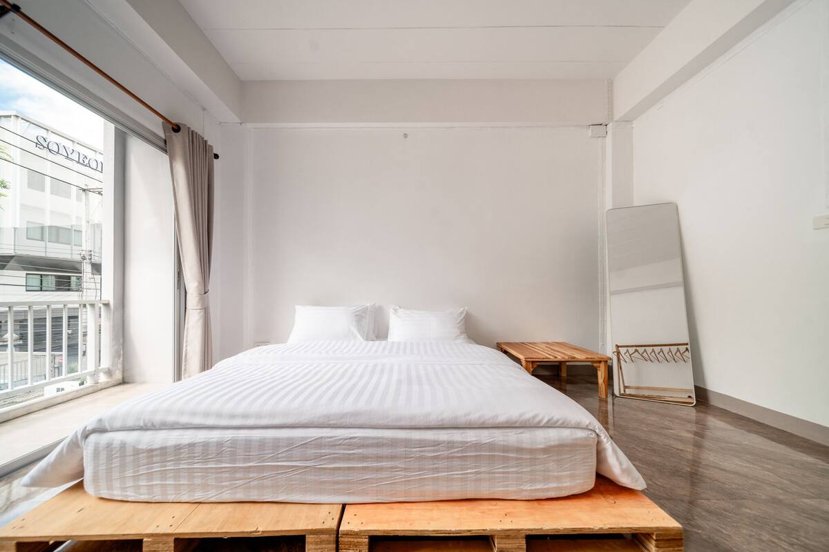 A minimalist bedroom with a large bed, a wooden nightstand, a full-length mirror, and a view of a building outside.