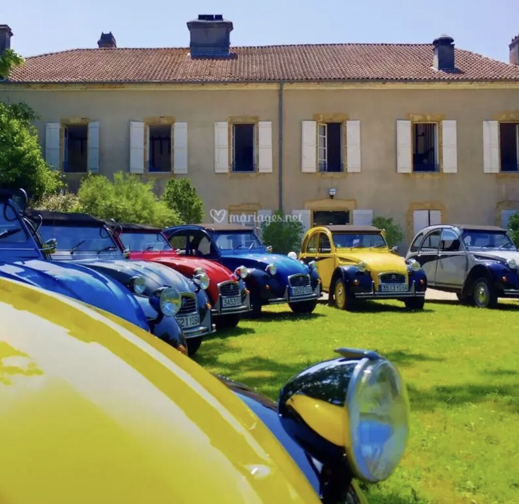 A row of vintage cars parked in front of a building with a tiled roof and white shutters.