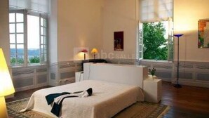 A bedroom with a large bed, a nightstand, a lamp, and a window with a view of greenery. - Grande Maison de Vacances (Projan)