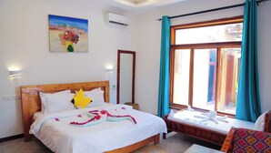 A hotel room with a wooden bed, a window with curtains, a painting on the wall, and a small table with a chair. - Palm Cultural Village (Paje)