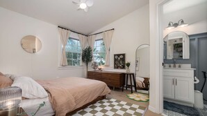 A bedroom with a bed, bedside table, dresser, and a desk with a chair. There is a window with curtains, a ceiling fan, and a mirror above the dresser.