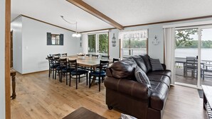 Een ruime woonkamer met een houten vloer, een leren bank, een eettafel met stoelen en grote ramen met uitzicht op het omliggende landschap.