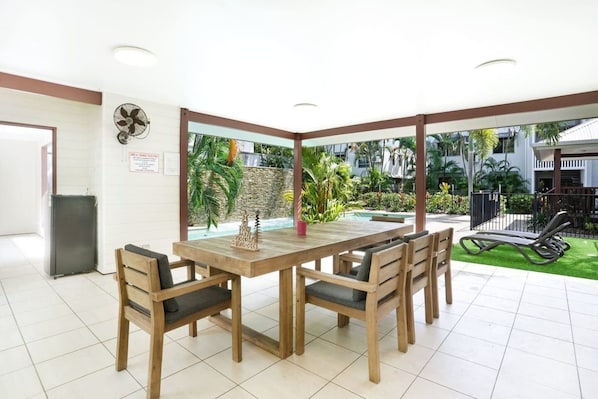 A spacious indoor dining area with a large wooden table and chairs, a view of a swimming pool and greenery outside, and a ceiling fan.