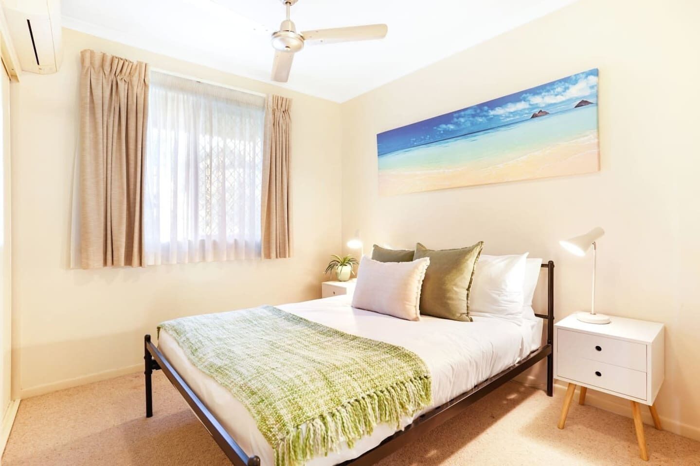 A bedroom with a bed, nightstands, a ceiling fan, a window with curtains, and a painting of a beach scene.