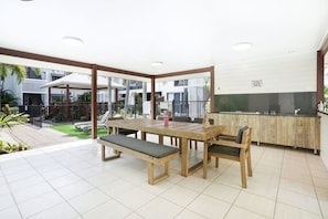 A spacious patio with a wooden table and benches, a kitchen area with a sink and countertop, and a view of a residential building in the background.