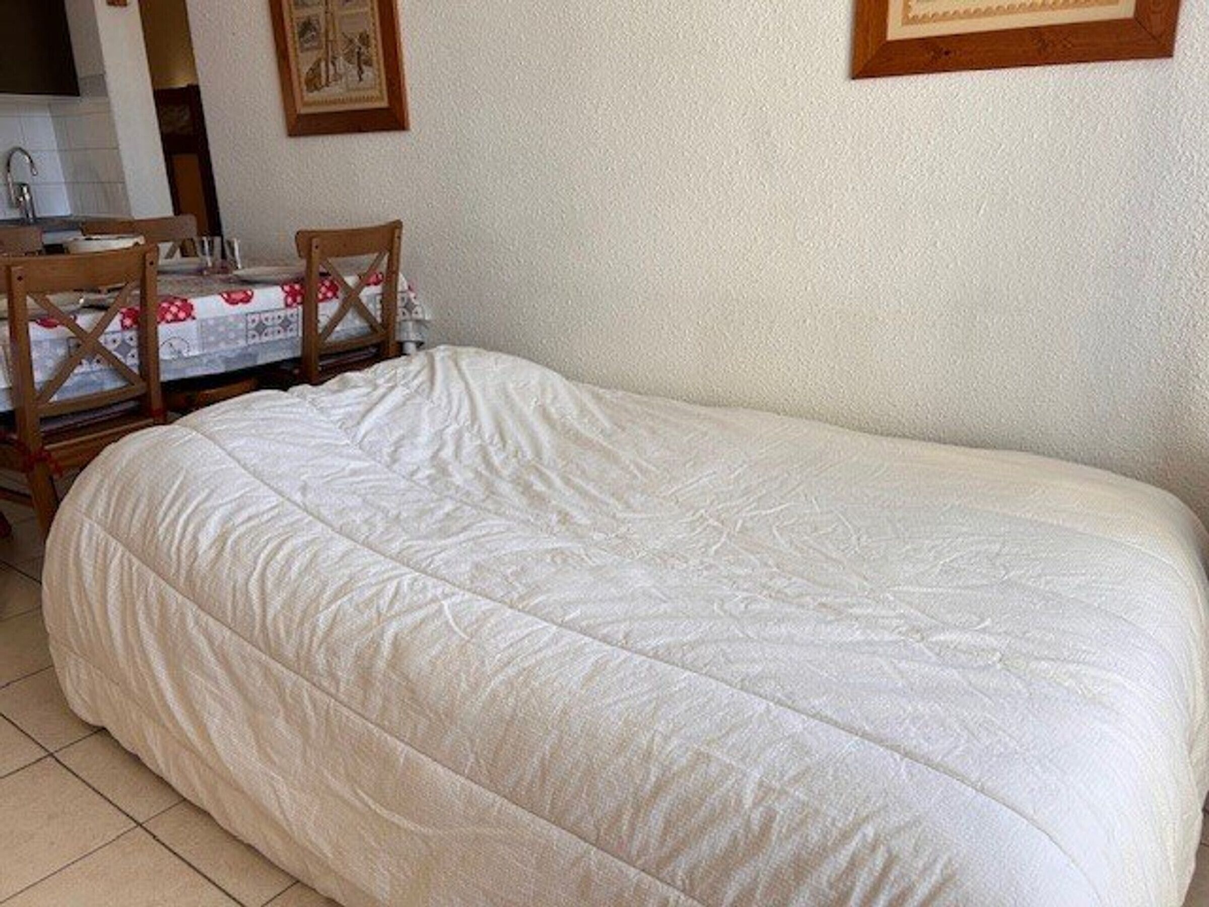 Un lit simple recouvert d’une couette blanche matelassée, dans une pièce dont le sol est carrelé.