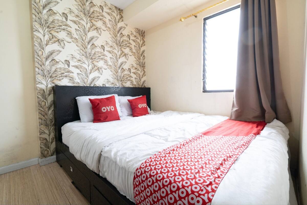 A neatly made bed with white and red bedding, a dark headboard, and a floral wallpaper pattern on the wall.