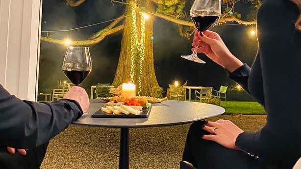 Dos personas sentadas en una mesa redonda al aire libre, con una vela y comida sobre la mesa, y un árbol grande al fondo.
