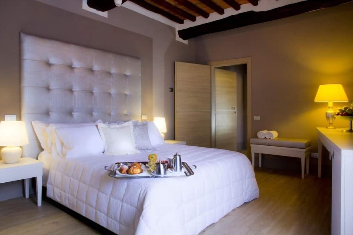A hotel room with a large bed, a bedside table with a lamp, a bench with towels, and a tray with breakfast items.