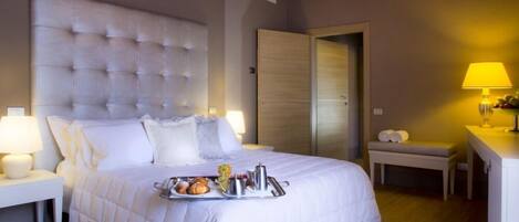 Una habitación de hotel con una cama grande, una mesita de noche con lámpara, un banco con toallas y una bandeja con alimentos para el desayuno.