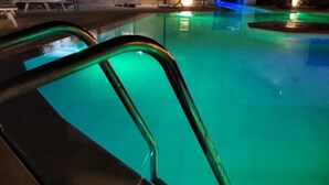 Una piscina con un borde iluminado en verde, una estructura de cristal al fondo y un banco cerca de la piscina.