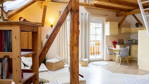 Un acogedor espacio tipo loft con vigas de madera a la vista, una cama en mezzanine, una pequeña cocina y una mesa de comedor con sillas.