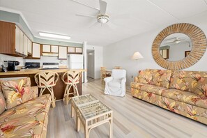 Uma sala de estar com sofá, cadeiras, mesa de centro, espelho redondo e área de cozinha com eletrodomésticos.
