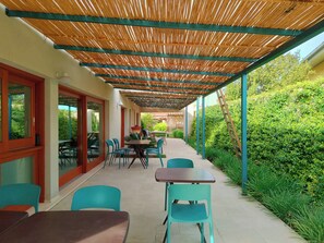 Une terrasse extérieure avec des tables et des chaises, un espace abrité avec un toit en bambou, et une vue sur la verdure.