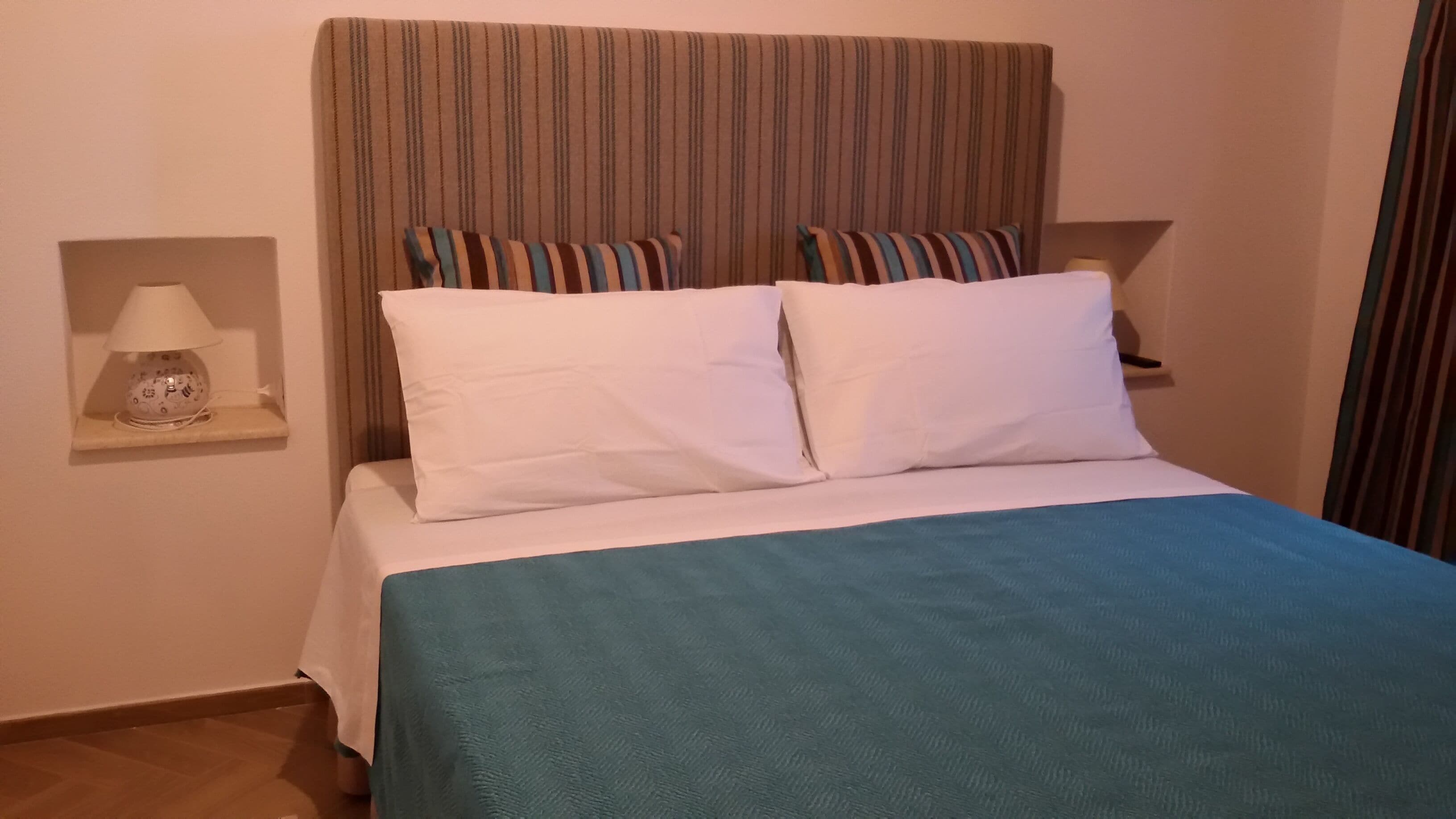 Un lit avec du linge de lit blanc, une couverture bleue et des oreillers à rayures. Il y a une table de chevet avec une lampe et un petit objet décoratif.