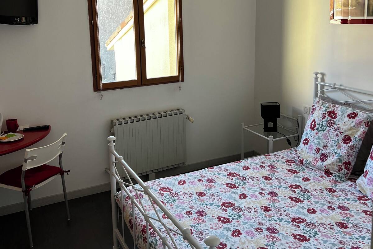 A bedroom with a bed, a desk, a chair, a radiator, a television, and a window.