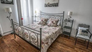 Un dormitorio con estructura de cama de metal, suelo de madera, colcha estampada y dos lámparas de noche, además de una silla con cojín.