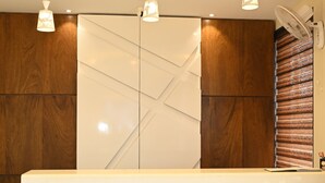 Balcão de recepção com painel de madeira ao fundo, iluminação pendente e um ventilador à direita.