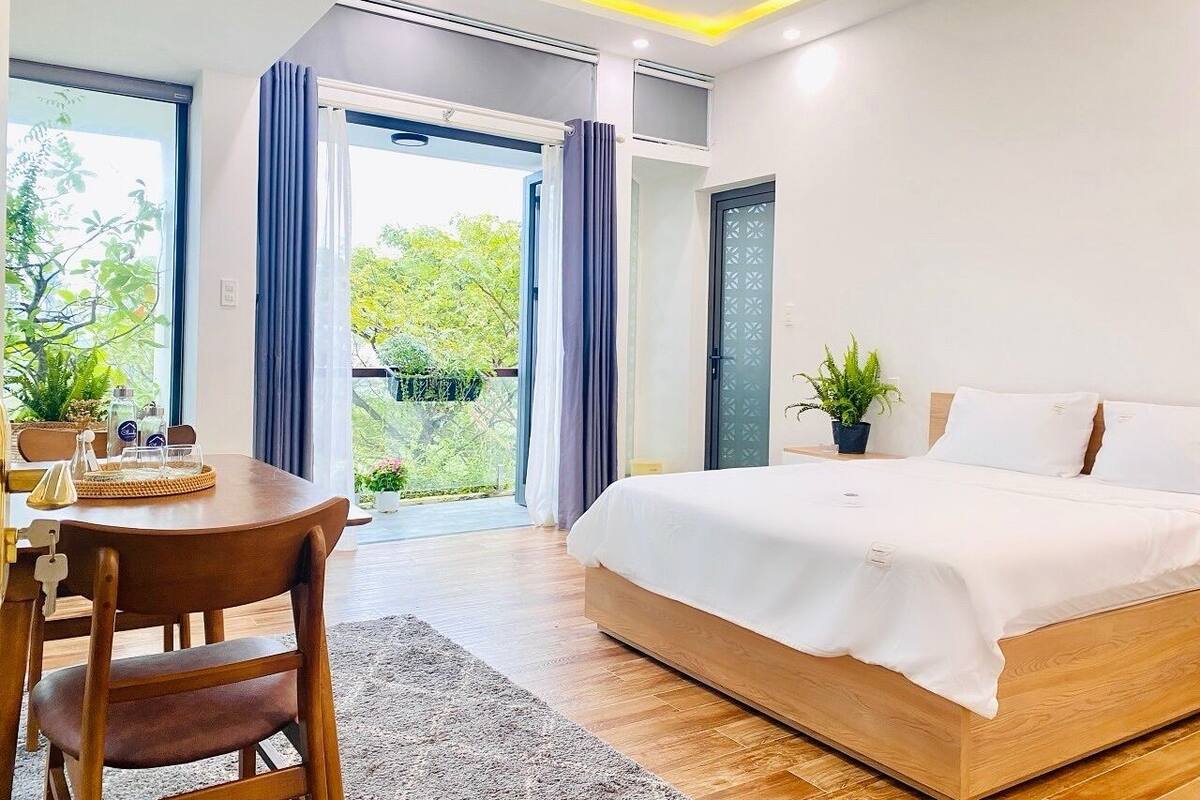 A modern bedroom with a large bed, a wooden desk, and a view of greenery outside.
