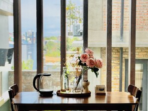 A table with a coffee pot, a vase with flowers, and a small card, set against a window with a view of buildings and greenery. - Cloudy Homestay Hoi An (Da Nang)