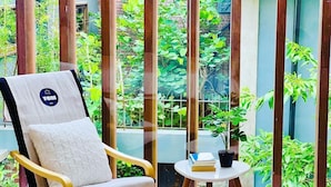 Una habitación con un ventanal grande, una silla de madera con cojín y una mesita con una planta y libros.