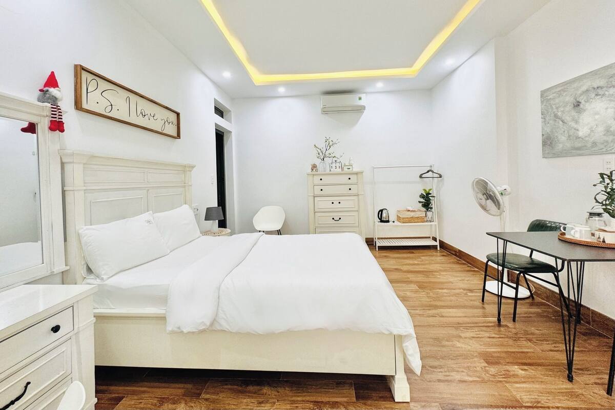 A modern bedroom with a bed, bedside tables, a dresser, a desk with a chair, a hanging plant, and a wall-mounted fan.