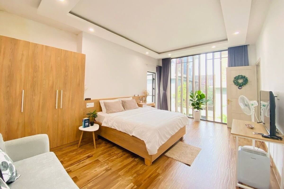 A modern bedroom with a wooden bed, a desk, a chair, and a large window with a view of greenery.