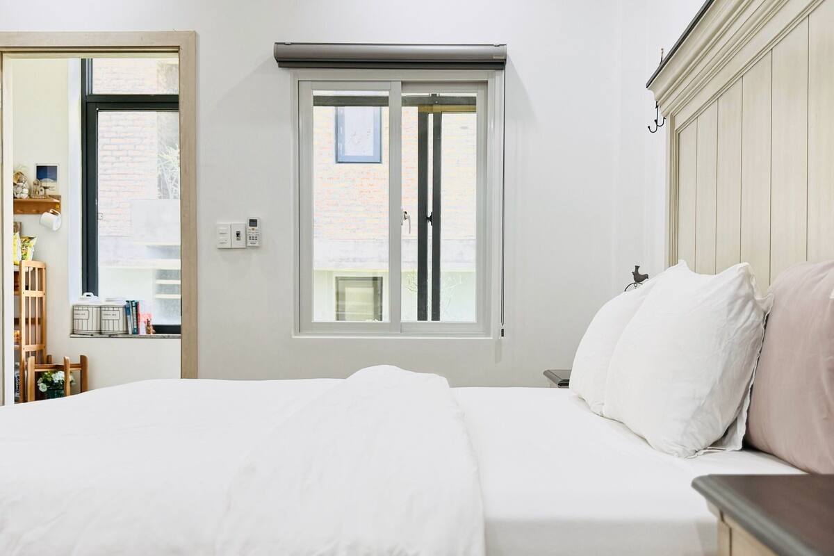 A bedroom with a bed, a window with blinds, a bedside table, and a wardrobe.