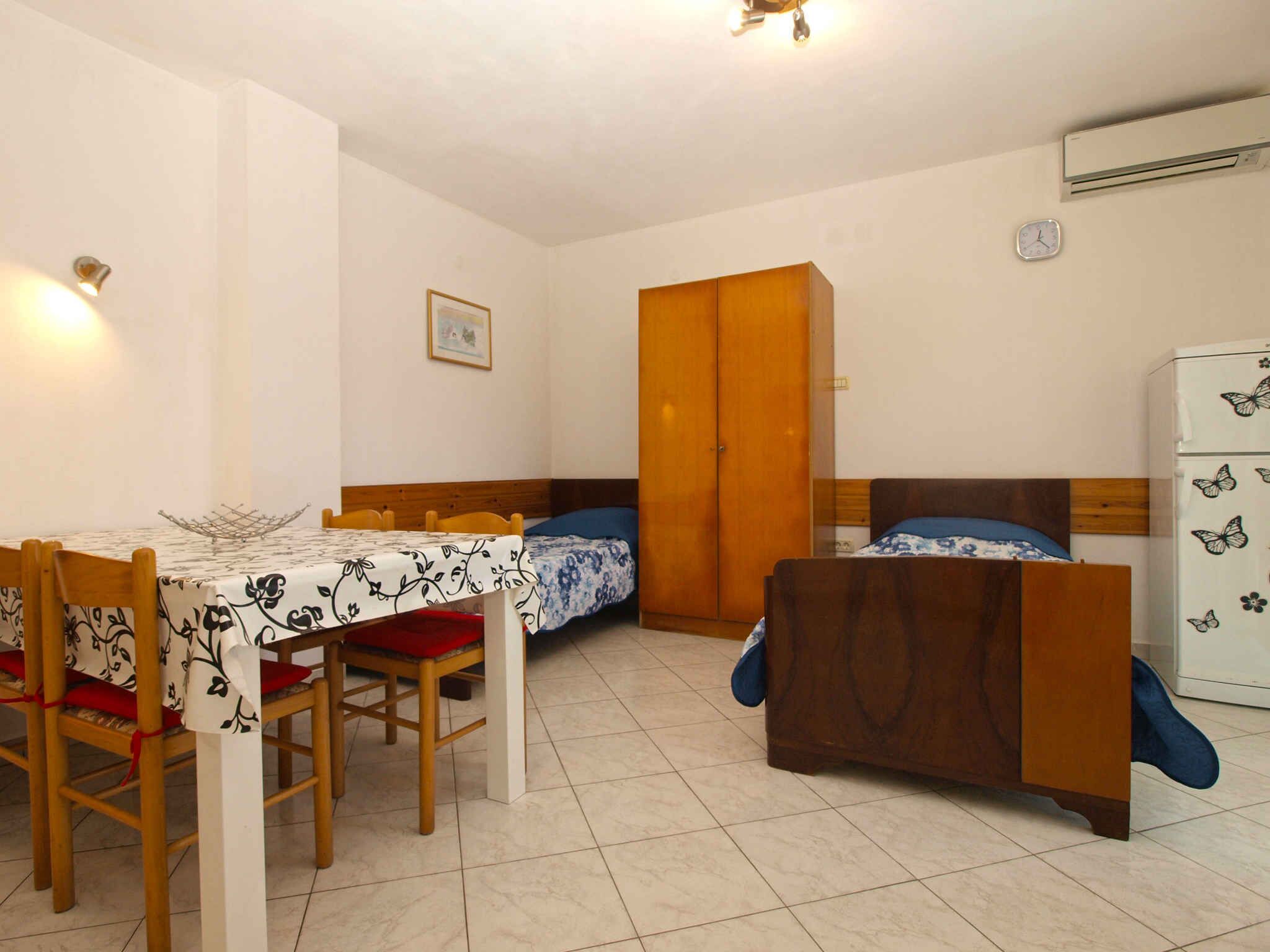 A small, well-lit room with a wooden dining table, two single beds, a wardrobe, and a refrigerator.