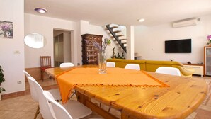 A dining area with a wooden table, white chairs, and an orange tablecloth. There is a staircase leading to an upper level, a TV mounted on the wall, and a cabinet with decorative items.