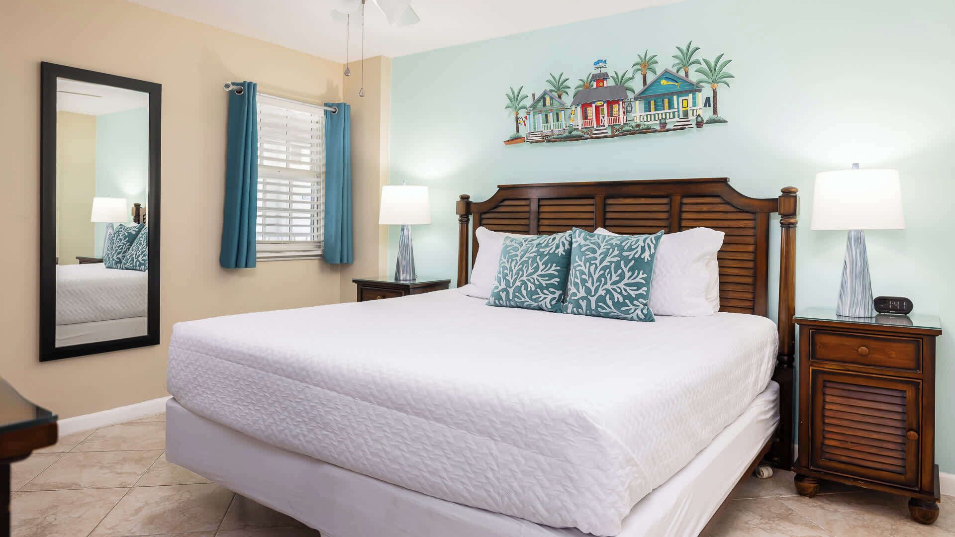 A hotel room with a large bed, a wooden headboard, a nightstand with a lamp, a mirror, and a wall with a tropical mural.