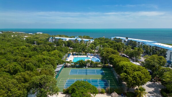 Vista aérea de un complejo turístico con cancha de tenis, piscina y vista al océano.