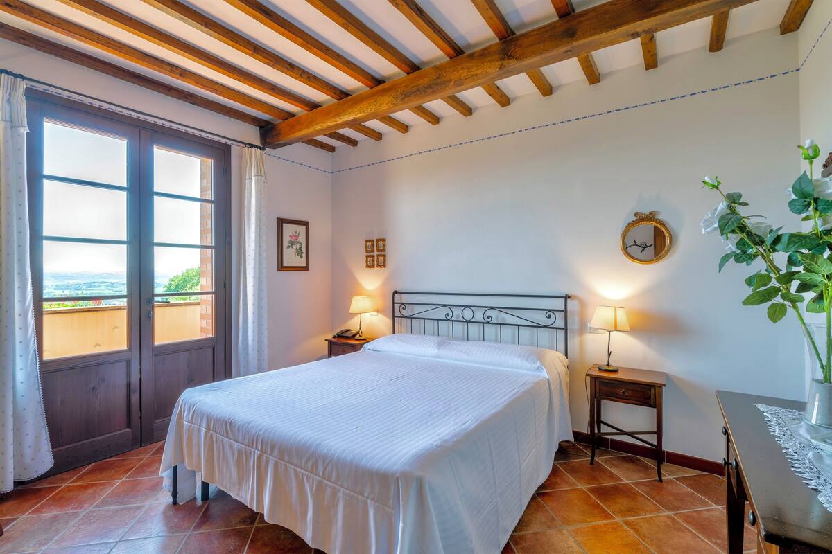 A bedroom with a bed, two bedside tables with lamps, a wall clock, and a view through a door.