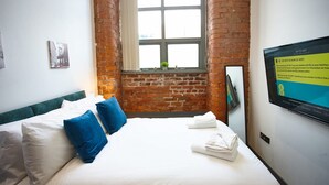 Ein Hotelzimmer mit Backsteinwand, einem großen Fenster, einem Bett mit weißer und blauer Bettwäsche und einem Fernseher, der ein WLAN-Symbol anzeigt.