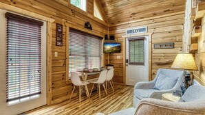 Interior acogedor de una cabaña con paredes de madera, un comedor con mesa y sillas, un sofá y un cartel que dice 'Bienvenido'.