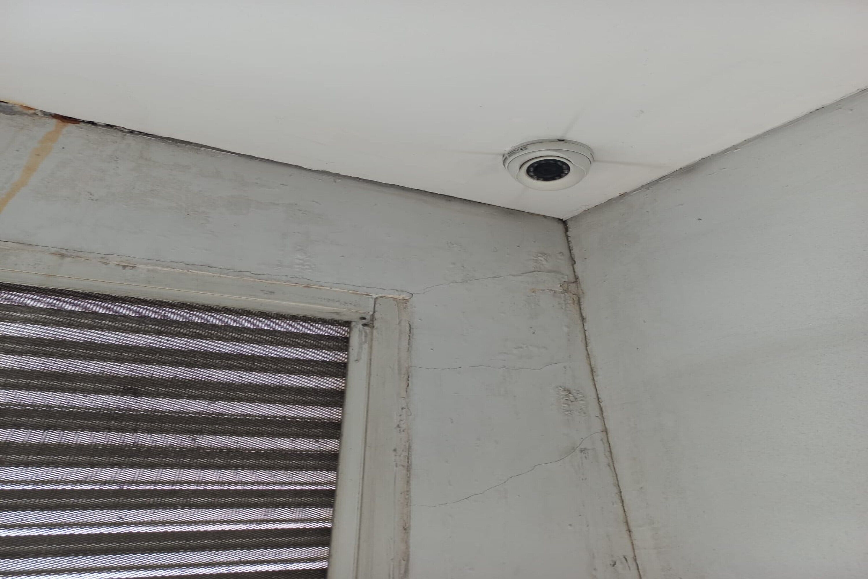 A corner of a room with a white ceiling, a window with horizontal blinds, and a visible crack in the wall.
